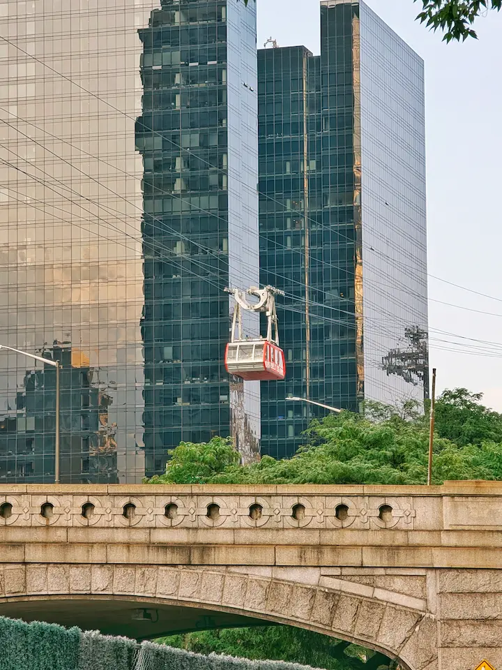 A red aerial tramway cain is suspended on cables, gilding between modern nyc skyscrapers and green trees